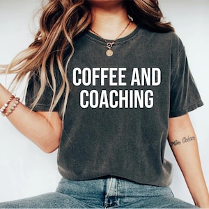 Puede incluir: Una camiseta gris oscura con el texto "COFFEE AND COACHING" impreso en blanco en la parte delantera. La camiseta la lleva puesta una persona con el pelo largo y casta&ntilde;o.