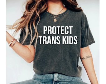 May include: A dark gray t-shirt with the text "PROTECT TRANS KIDS" printed in white on the front. The shirt is worn by a person with long brown hair and is paired with blue jeans.