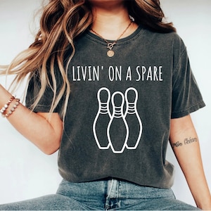 May include: Dark gray t-shirt with the text "Livin' on a Spare" above a graphic of three white bowling pins. The shirt has a vintage, worn look.