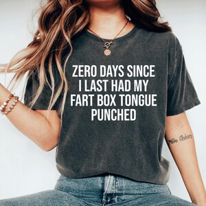 May include: A charcoal gray short-sleeve t-shirt featuring the white text "ZERO DAYS SINCE I LAST HAD MY FART BOX TONGUE PUNCHED" printed in a bold, sans-serif font across the center of the chest. The garment has a crew neckline and a relaxed fit.