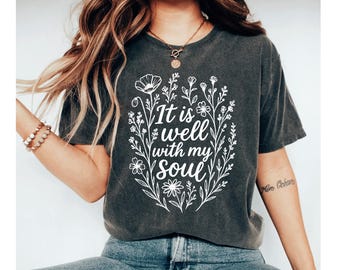 May include: Dark gray t-shirt with the text "It is well with my soul" in a white floral design. The design features various flowers and leaves, creating a decorative border around the text. The shirt is a casual style.