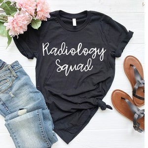 May include: Black t-shirt with white text that reads "Radiology Squad". The shirt is shown on a white wooden surface with a pair of brown sandals and blue jeans.