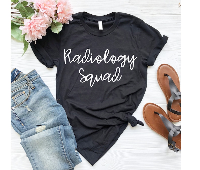 May include: Black t-shirt with white text that reads "Radiology Squad". The shirt is shown on a white wooden surface with a pair of brown sandals and blue jeans.
