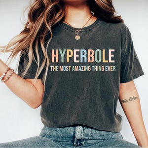 May include: A dark gray t-shirt with the text "HYPERBOLE THE MOST AMAZING THING EVER" in rainbow colors.