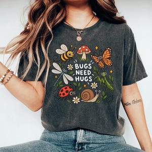 May include: A charcoal gray t-shirt featuring a colorful graphic of a bee, butterfly, dragonfly, ladybug, snail, praying mantis, and mushroom with the text "BUGS NEED HUGS" surrounded by small white flowers and yellow star accents.