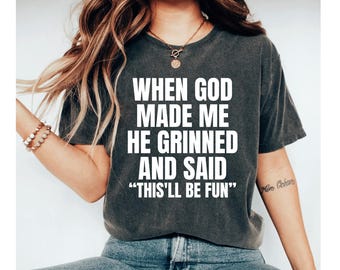May include: Dark gray t-shirt with the white text: "WHEN GOD MADE ME HE GRINNED AND SAID 'THIS'LL BE FUN'". The shirt is a crew neck style. The model is wearing blue jeans and a bracelet.
