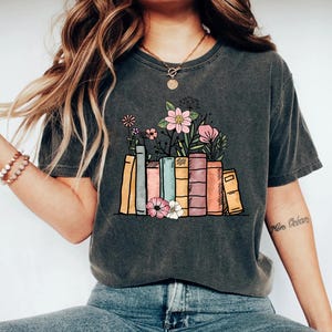 May include: A dark gray t-shirt with a graphic of a stack of colorful books with flowers growing out of the top. The text "More Books" is written on the right sleeve.