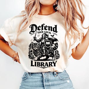 May include: Cream-colored t-shirt with a black graphic. The graphic features a knight in armor holding a sword, riding a cat, with the words "Defend Your Local Library" above and below the image.