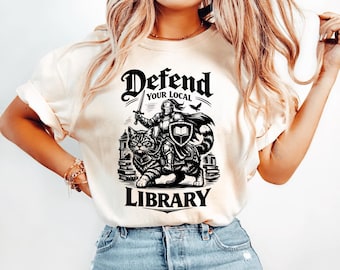 May include: Cream-colored t-shirt with a black graphic. The graphic features a knight in armor holding a sword, riding a cat, with the words "Defend Your Local Library" above and below the image.