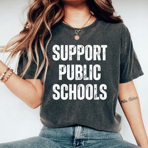 May include: Dark gray t-shirt with the words "SUPPORT PUBLIC SCHOOLS" in large white letters. The shirt is a crew neck style and appears to be made of a soft material. The person is wearing blue jeans.