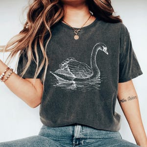 May include: Dark gray t-shirt featuring a white illustration of a swan swimming on water. The swan is detailed with visible feathers and a curved neck. The shirt has a vintage, worn look.