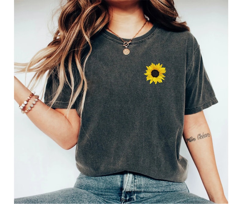 May include: A dark gray t-shirt with a yellow sunflower graphic on the left chest. The shirt has a relaxed fit and a crew neck.