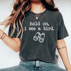 May include: Dark grey t-shirt with the text "hold on, i see a bird." and a white graphic of binoculars. The shirt is a casual style, suitable for everyday wear.
