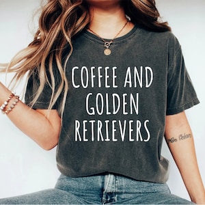 May include: Dark gray t-shirt with the words "COFFEE AND GOLDEN RETRIEVERS" in white block letters. The shirt is worn by a person with long blonde hair and a bracelet on their wrist.