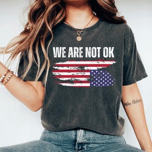 May include: A dark gray t-shirt with a distressed American flag design and the text "WE ARE NOT OK" in white.