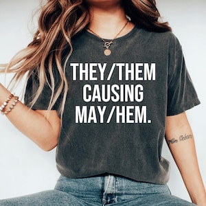 May include: A dark gray t-shirt with white text that reads "THEY/THEM CAUSING MAY/HEM."
