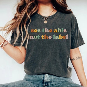May include: A dark grey t-shirt with the text "see the able not the label" in colourful letters. The letters are in a retro font style.