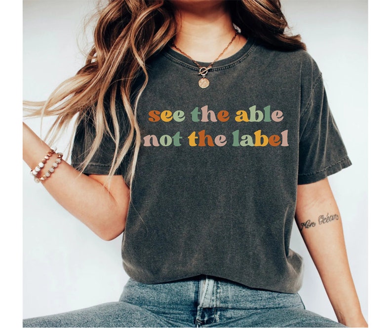 May include: A dark grey t-shirt with the text "see the able not the label" in colourful letters. The letters are in a retro font style.