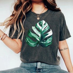 May include: A dark gray t-shirt with a green and white monstera leaf print. The shirt has a relaxed fit and a crew neck.