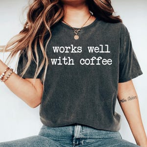 May include: Dark gray t-shirt with the white text "works well with coffee". The shirt is a short-sleeved, crew neck style. The shirt is paired with blue jeans. The overall theme is casual wear.