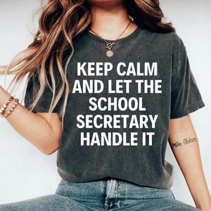 May include: A dark gray t-shirt with the text "Keep Calm and Let the School Secretary Handle It" printed in white.