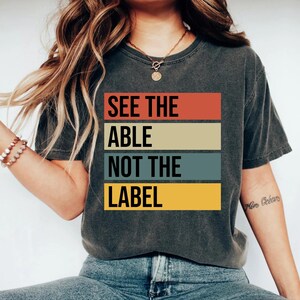 Op de afbeelding: Donkergrijs t-shirt met de tekst "SEE THE ABLE NOT THE LABEL" in een retro kleurblokontwerp. Het shirt heeft een ronde hals en korte mouwen. Het shirt is gemaakt van een zacht materiaal.
