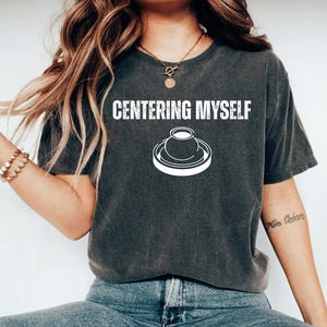 May include: Dark gray t-shirt with the text "CENTERING MYSELF" above a white graphic of a pottery wheel and a pot. The shirt is a comfortable, casual style.