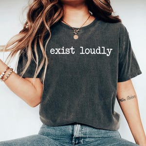 May include: A dark gray t-shirt with the text "exist loudly" printed in white on the front. The shirt is worn by a person with long brown hair and a necklace.