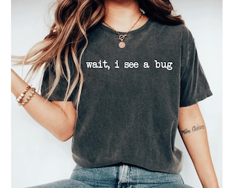 May include: Dark gray t-shirt with the white text "wait, i see a bug". The shirt has short sleeves and a crew neck. The shirt is worn with blue jeans.