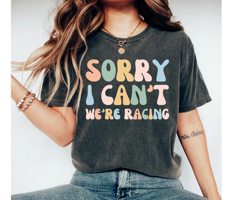 Op de afbeelding: Een donkergrijs T-shirt met een kleurrijke afbeelding met de tekst "Sorry, ik kan niet, we racen".