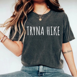 May include: A dark gray t-shirt with the text "TRYNA HIKE" printed in white on the front. The shirt is worn by a person with long brown hair and a gold necklace.