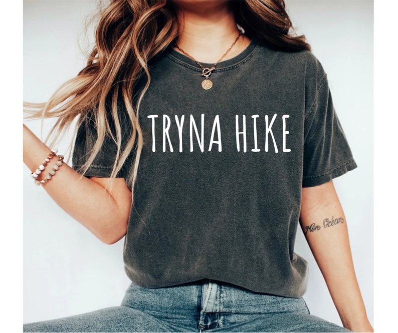 May include: A dark gray t-shirt with the text "TRYNA HIKE" printed in white on the front. The shirt is worn by a person with long brown hair and a gold necklace.