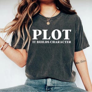 May include: A dark gray t-shirt with white text that reads "PLOT IT BUILDS CHARACTER".