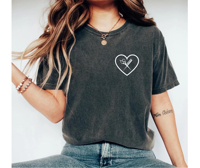 K&ouml;nnte beinhalten: Ein dunkelgraues T-Shirt mit einem wei&szlig;en Herzdesign, das eine Schere und einen Kamm im Herzen zeigt.