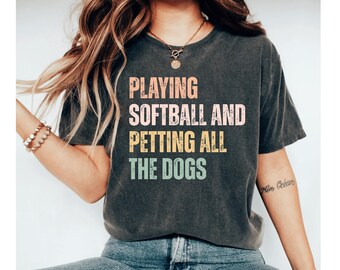 May include: Dark gray t-shirt with the text "PLAYING SOFTBALL AND PETTING ALL THE DOGS" in a colorful, retro font. The shirt is a casual style, suitable for everyday wear.