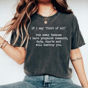 May include: A charcoal gray short-sleeve t-shirt featuring white text that reads: if i say "first of all" run away because i have prepared research, data, charts and will destroy you. The shirt has a crew neckline and a relaxed fit.