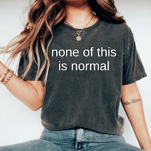 May include: Dark gray t-shirt with the text "none of this is normal" in white. The shirt is a short-sleeved, crew neck style. The model is wearing blue jeans and a gold necklace.
