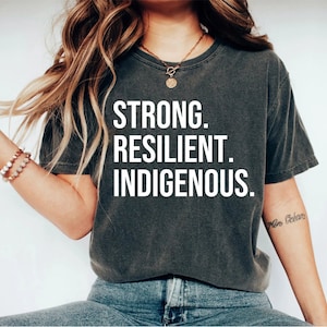 May include: A dark gray t-shirt with the text "STRONG. RESILIENT. INDIGENOUS." printed in white.