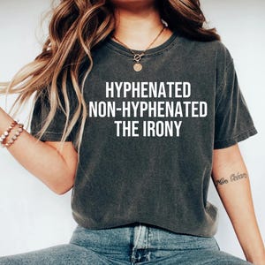 K&ouml;nnte beinhalten: Ein dunkelgraues T-Shirt mit dem Text "HYPHENATED NON-HYPHENATED THE IRONY" in wei&szlig; gedruckt.