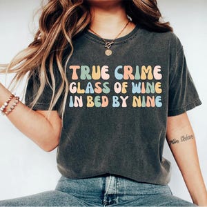 May include: A dark gray t-shirt with the text "TRUE CRIME GLASS OF WINE IN BED BY NINE" in colorful, retro-style lettering.