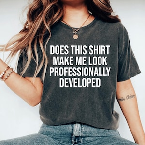 May include: A charcoal gray short-sleeve t-shirt featuring the white text DOES THIS SHIRT MAKE ME LOOK PROFESSIONALLY DEVELOPED printed in a bold, sans-serif font across the center of the chest. The garment has a crew neckline and a relaxed fit.