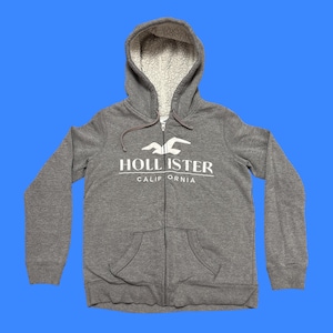 May include: Gray zip-up hoodie with a white sherpa-lined hood. The hoodie features a white logo that says "HOLLISTER CALIFORNIA" and two front pockets. The background is a solid blue.