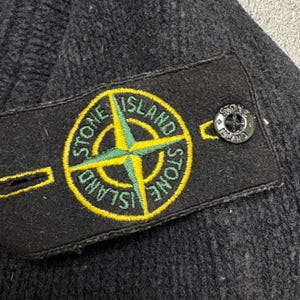 Op de afbeelding: Close-up van een donkergrijze gebreide beanie met een zwart label met een geel en groen kompasroosontwerp en de woorden "STONE ISLAND". Een kleine, zwarte knoop met de merknaam is zichtbaar.