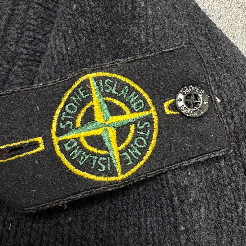 Op de afbeelding: Close-up van een donkergrijze gebreide beanie met een zwart label met een geel en groen kompasroosontwerp en de woorden "STONE ISLAND". Een kleine, zwarte knoop met de merknaam is zichtbaar.