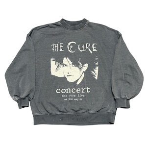 Op de afbeelding: Een donkergrijze sweatshirt met een grafische print van The Cure. Het ontwerp bevat de bandnaam, een portret en het woord "concert" met "the cure live uk tour may 84" eronder gedrukt. De sweatshirt heeft een ronde hals en lange mouwen.