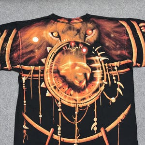 May include: Black t-shirt featuring a detailed graphic of a lion's face and a circular design with a lion's head. The design is in shades of orange and gold, with intricate details. The shirt has a crew neck.