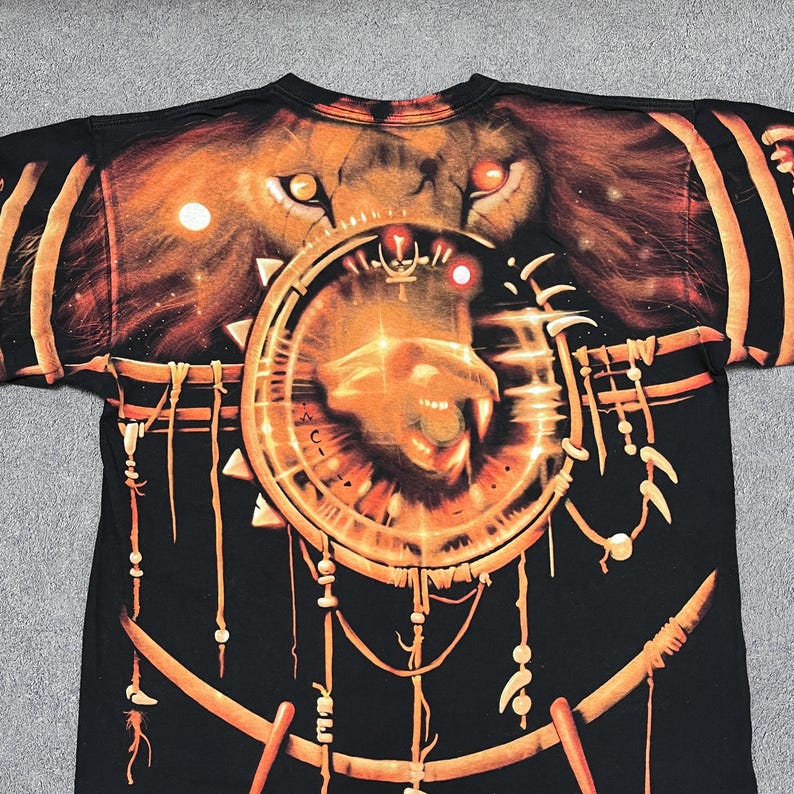 May include: Black t-shirt featuring a detailed graphic of a lion's face and a circular design with a lion's head. The design is in shades of orange and gold, with intricate details. The shirt has a crew neck.