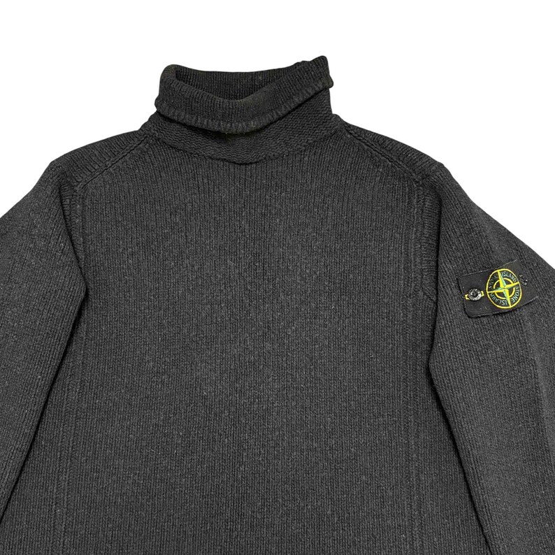 Op de afbeelding: Een donkergrijze coltrui met een geribbelde brei textuur. De trui heeft een hoge, opgevouwen kraag en een kleine, vierkante patch met een geel en groen logo op de linkermouw.