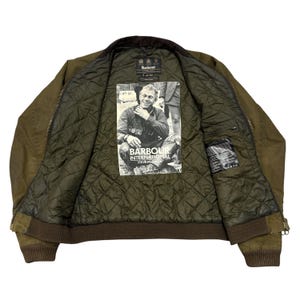 May include: An olive green jacket with a dark brown ribbed collar and cuffs. The interior features a quilted lining and a large graphic print of Steve McQueen with the text Barbour International Steve McQueen. A brand label is at the neck.