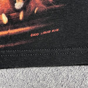 May include: Close-up of a black t-shirt with a graphic design. The text "&copy;2001 LIQUID BLUE" is printed in red on the lower right side of the shirt. The shirt has a double-stitched hem.